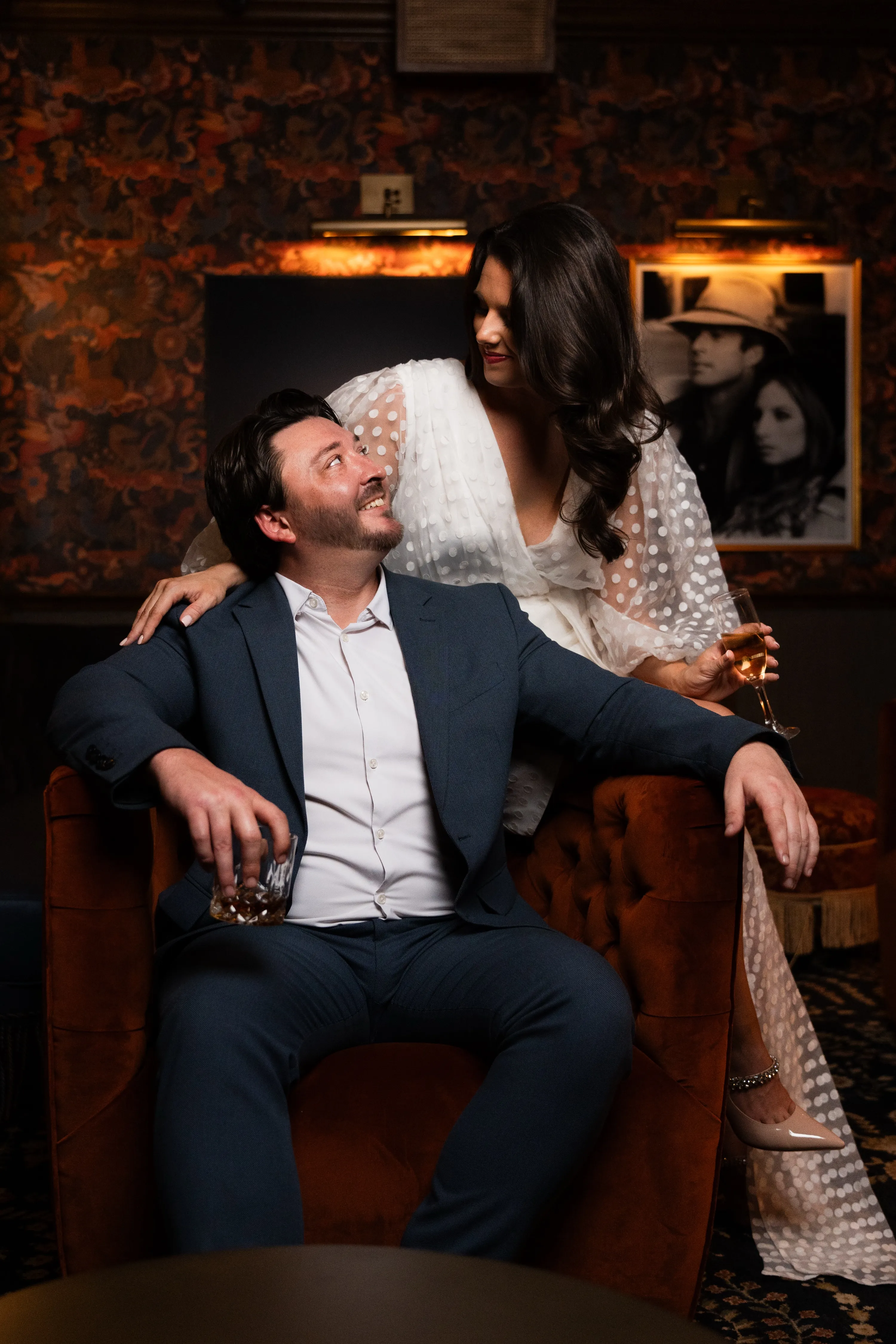 Pareja comprometida mirándose en un sillón de terciopelo con champaña durante su sesión de pareja en el bar de Columbian Country Club