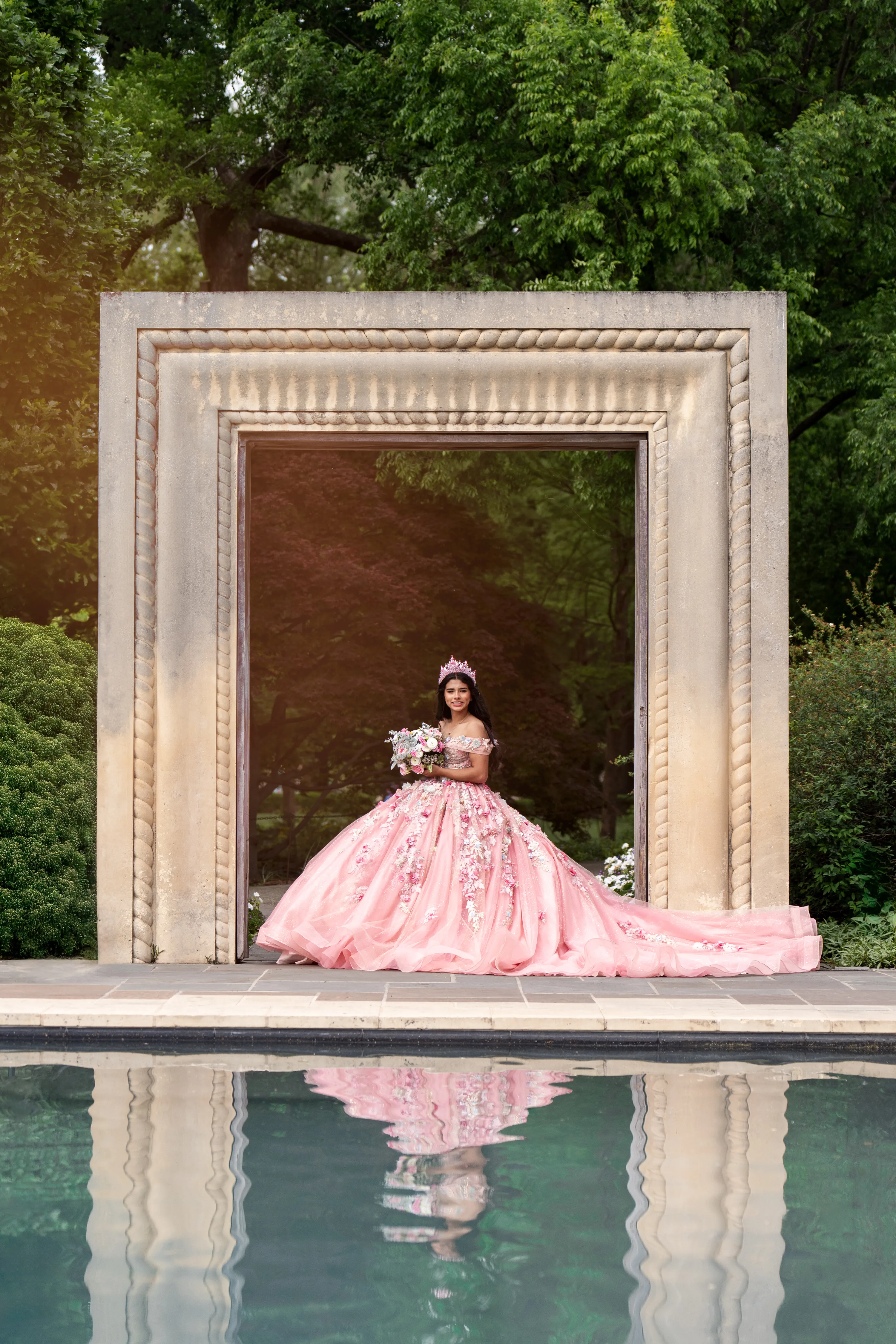 Quinceañera con vestido de quince rosa floral sentada en un arco de piedra sobre un estanque en el Dallas Arboretum