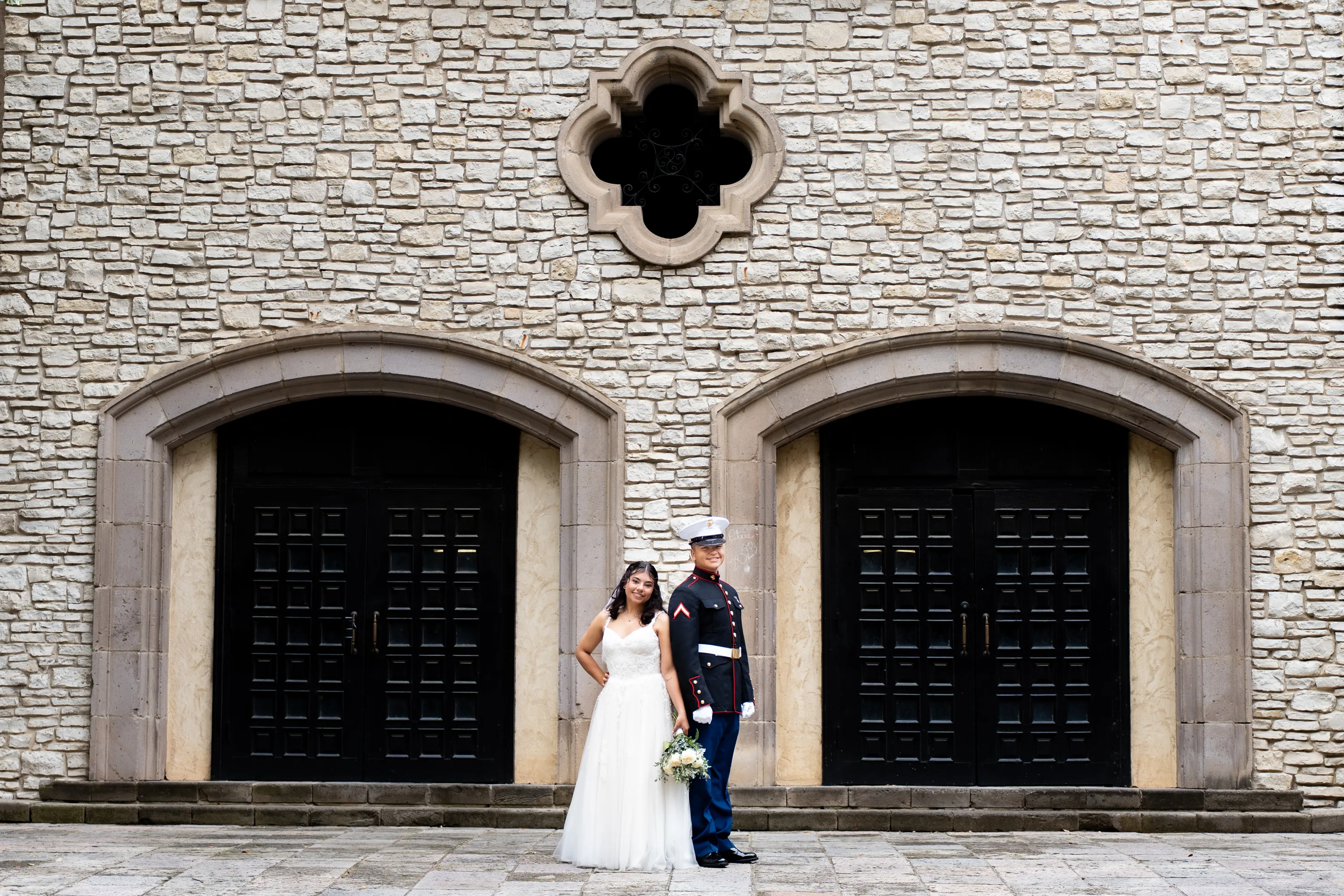 Novia y novio marine con uniforme de gala frente a puertas de arco de piedra en el Mandalay Canal de Irving, Texas