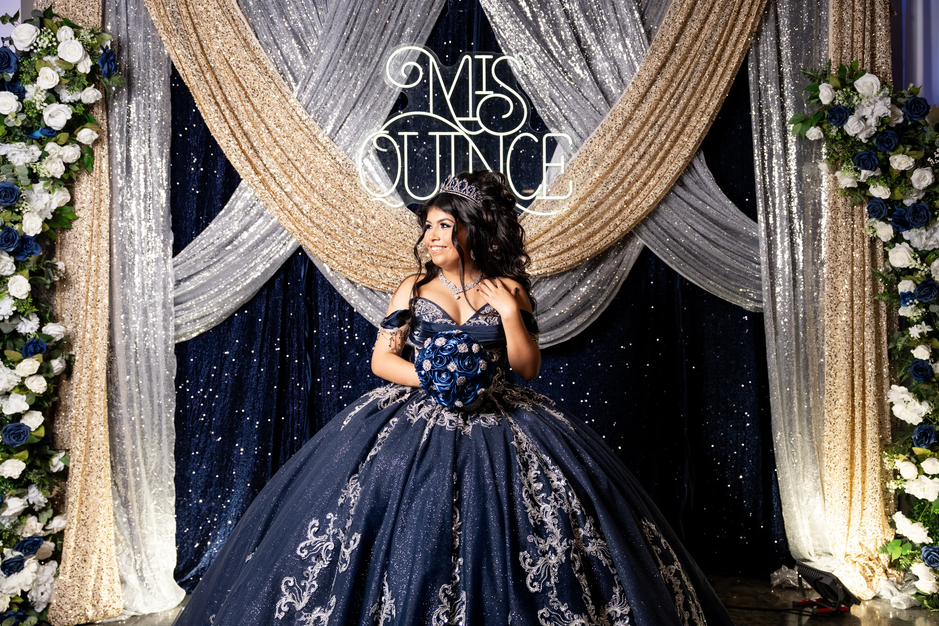 Quinceañera con vestido azul marino y rosas azules frente a un letrero de Mis Quince en Diamond Ranch Event Center en Waxahachie, Texas