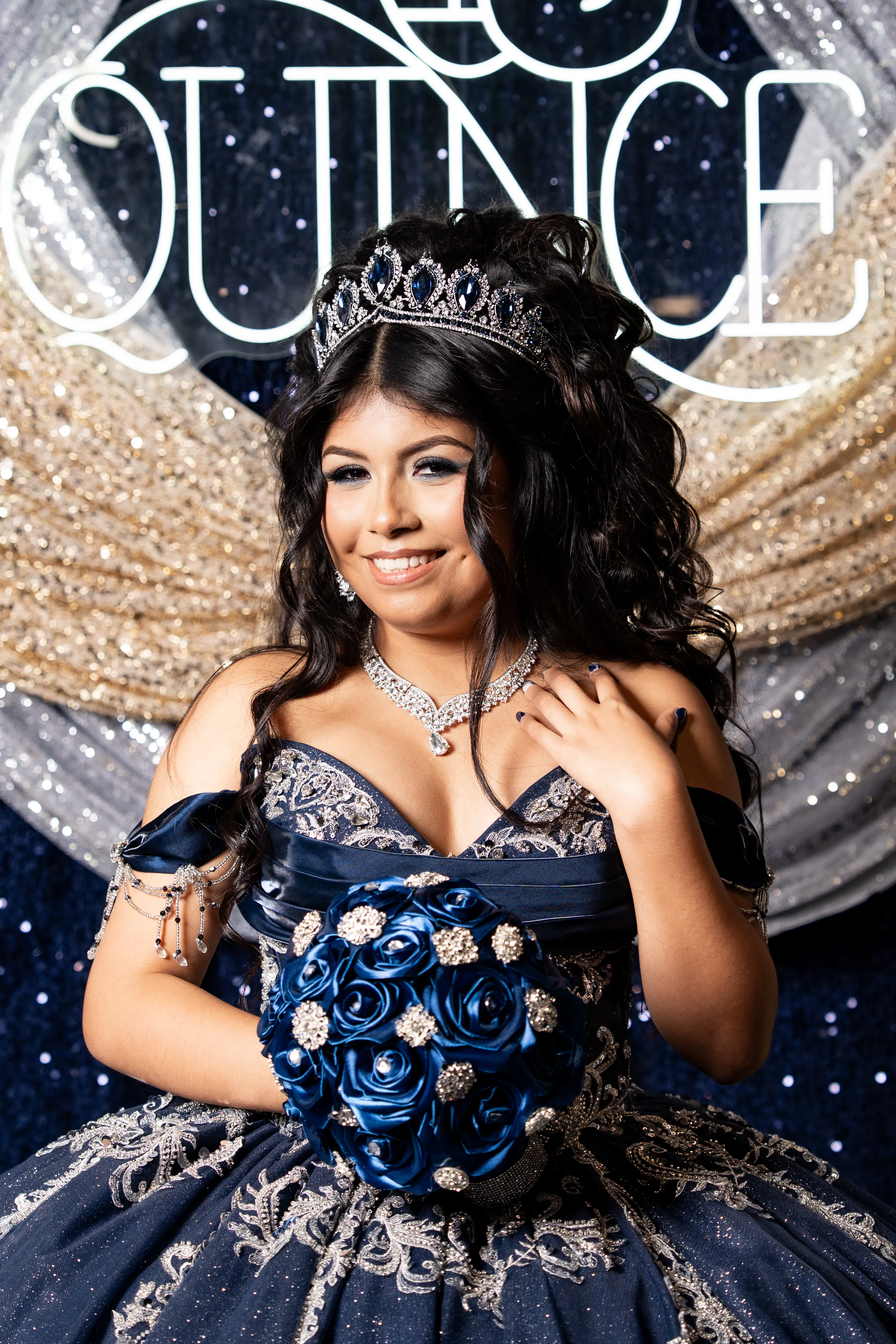 Quinceañera con vestido de quince azul marino y tiara sosteniendo rosas azules bajo un letrero de Mis Quince en Diamond Ranch Event Center en Waxahachie, Texas
