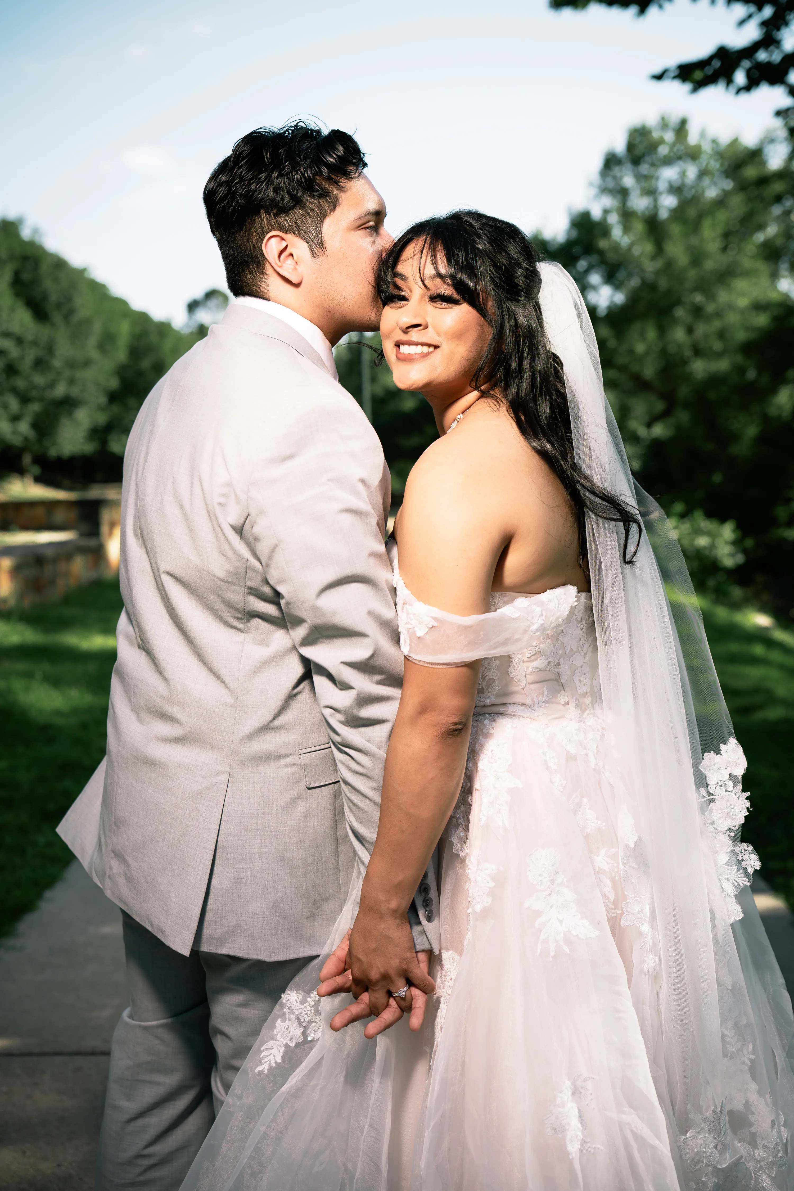 Novio besando a su novia sonriente con velo en un sendero de Turtle Creek Park en Dallas