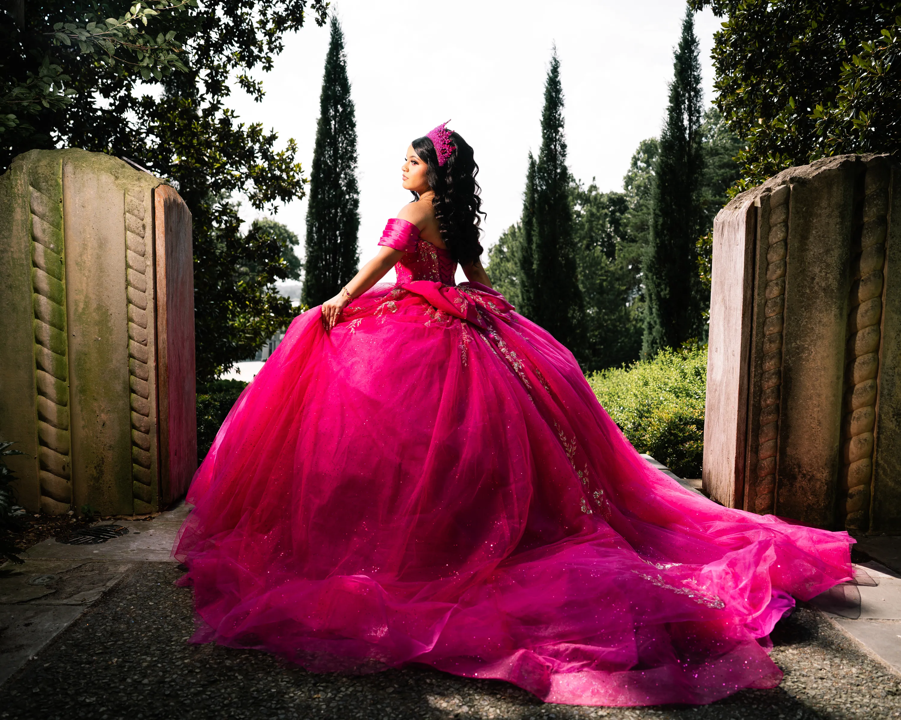 Quinceañera con vestido fucsia y corona caminando entre las puertas de piedra del jardín del Dallas Arboretum