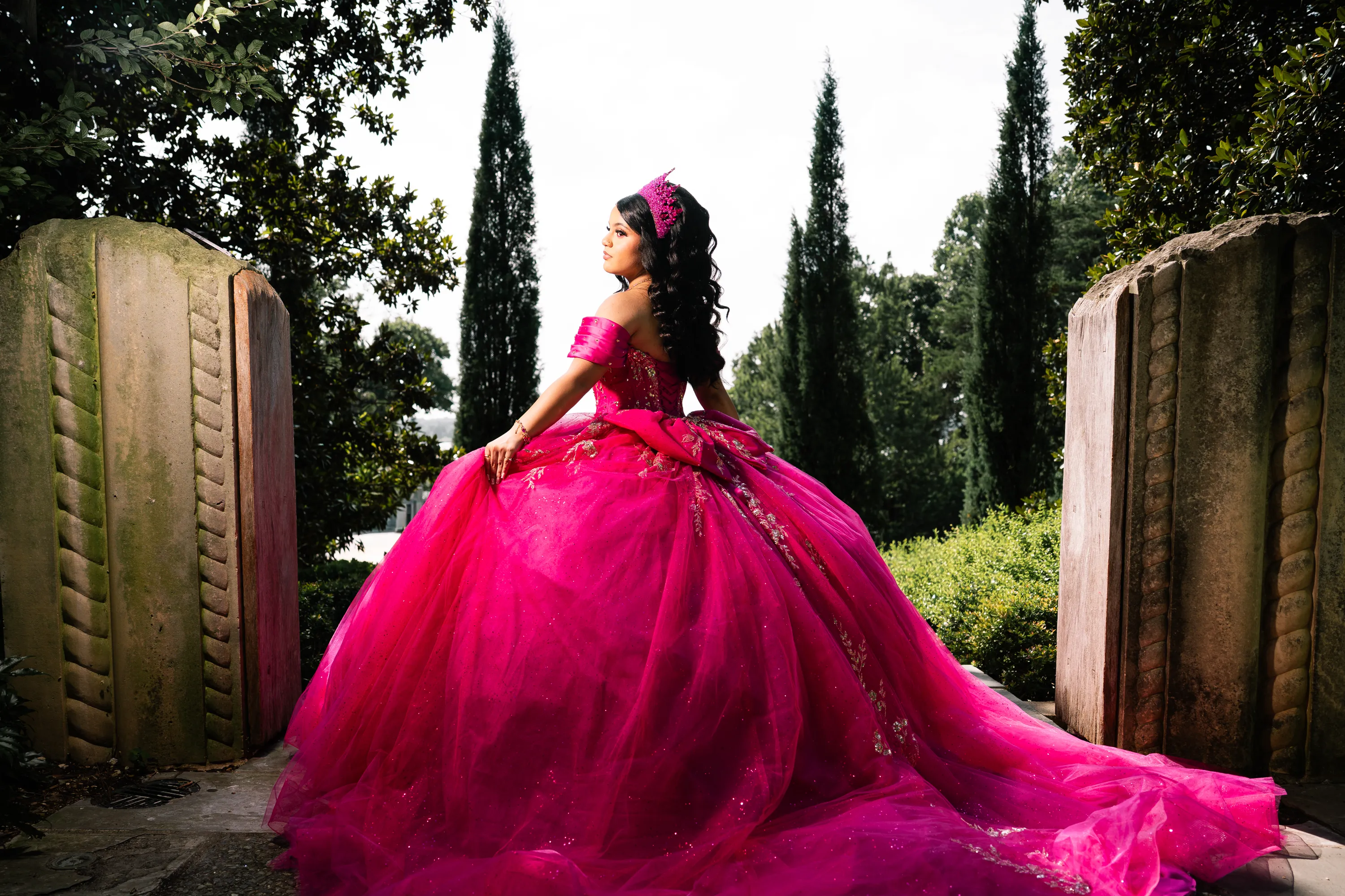 Quinceañera in a fuchsia quince gown and crown walking between stone garden gates at the Dallas Arboretum
