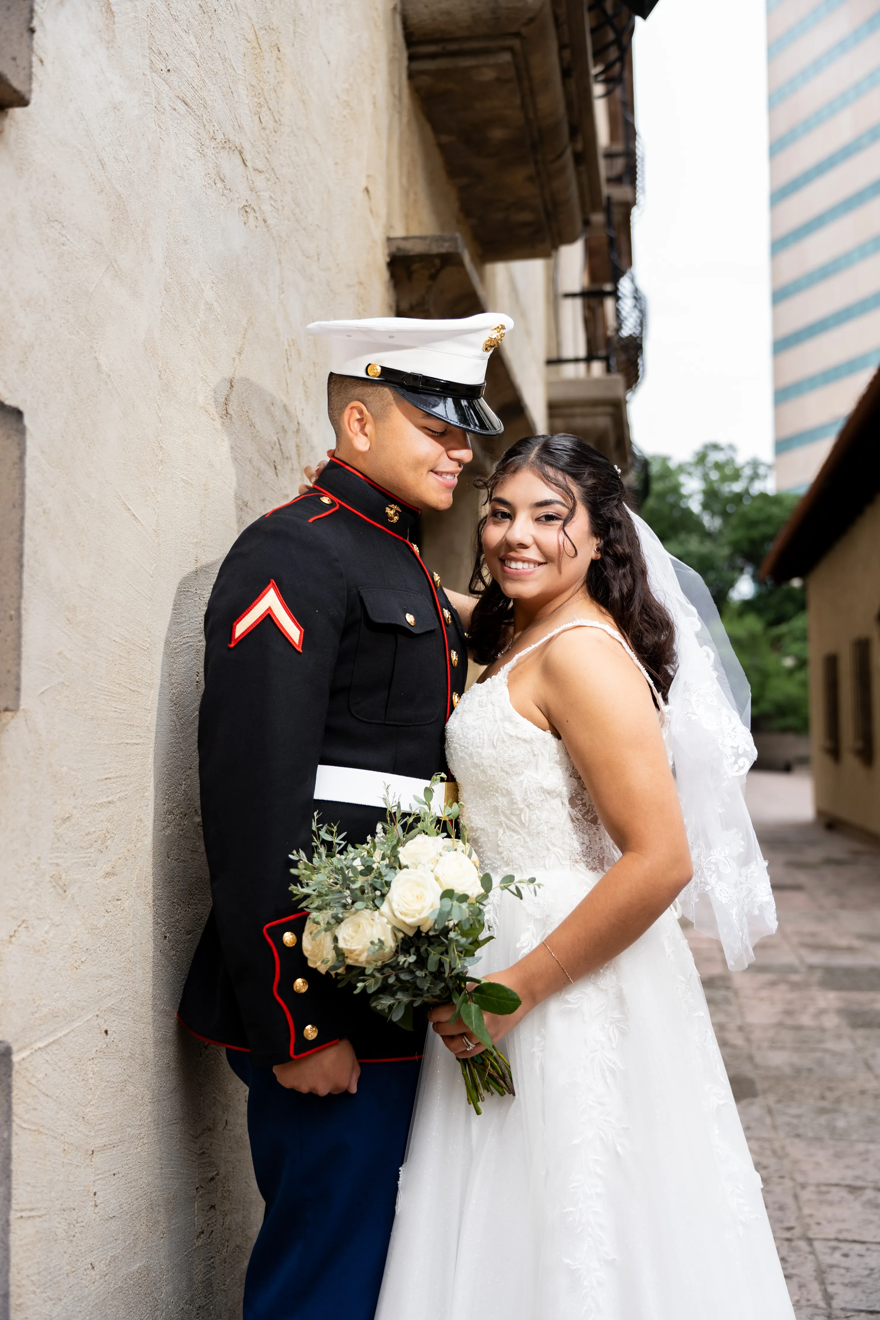 Novia sonriente con un ramo de rosas blancas junto a su novio marine con uniforme de gala en el Mandalay Canal de Irving, Texas