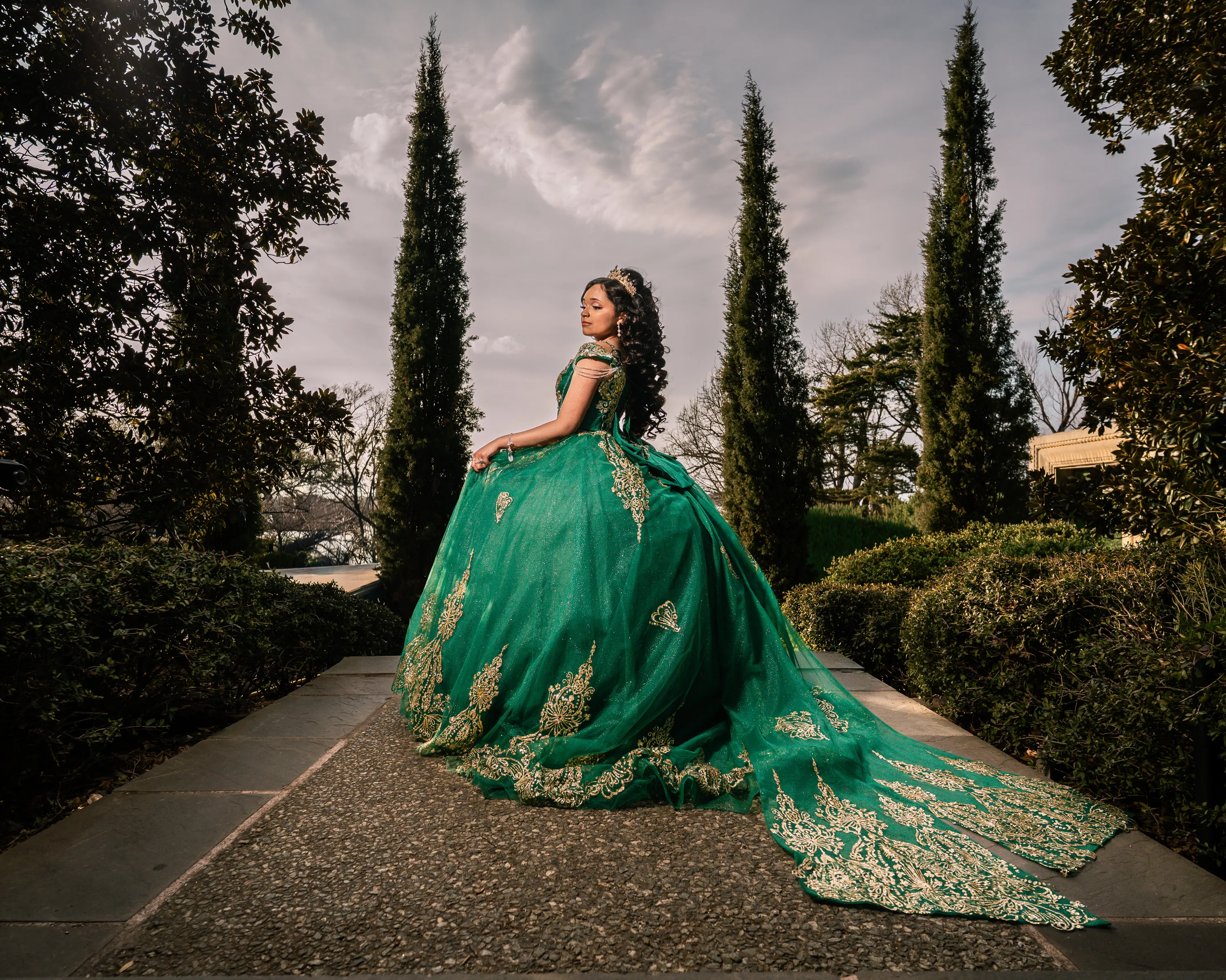 Quinceañera con vestido verde esmeralda y dorado en un sendero del jardín del Dallas Arboretum