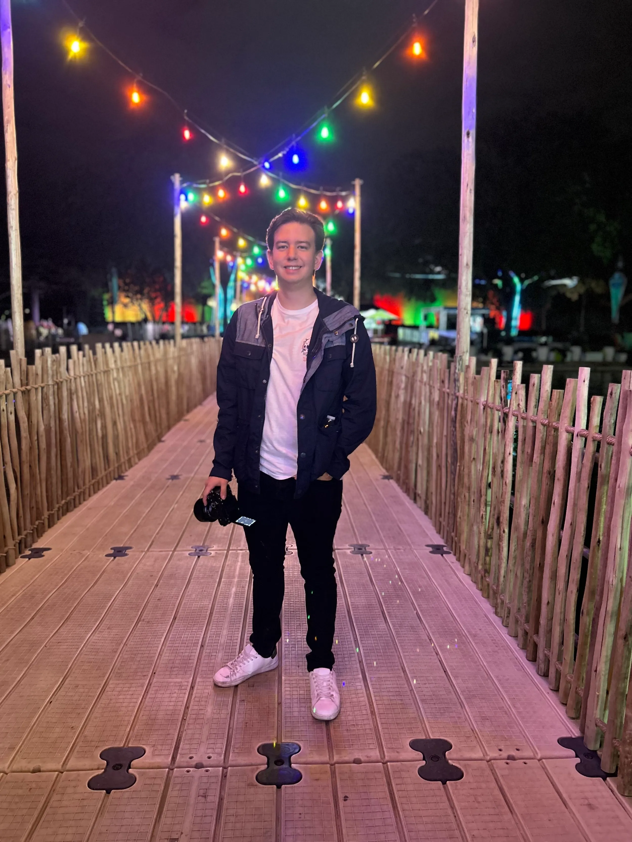 Un joven fotógrafo profesional de pie en un camino de madera iluminado con luces de colores de noche, sosteniendo una cámara y sonriendo.