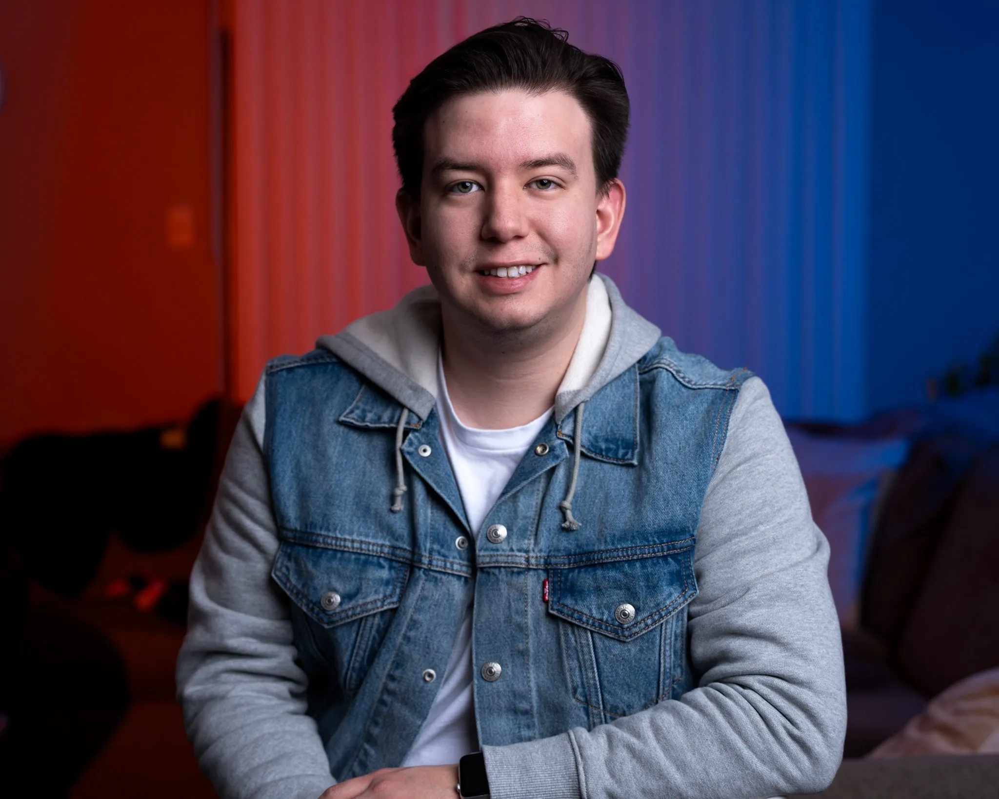 El fotógrafo profesional de quinceañeras Rony Lanza sentado en interiores, sonriendo, con una sudadera de mezclilla y gris, con luces rojas y azules al fondo.