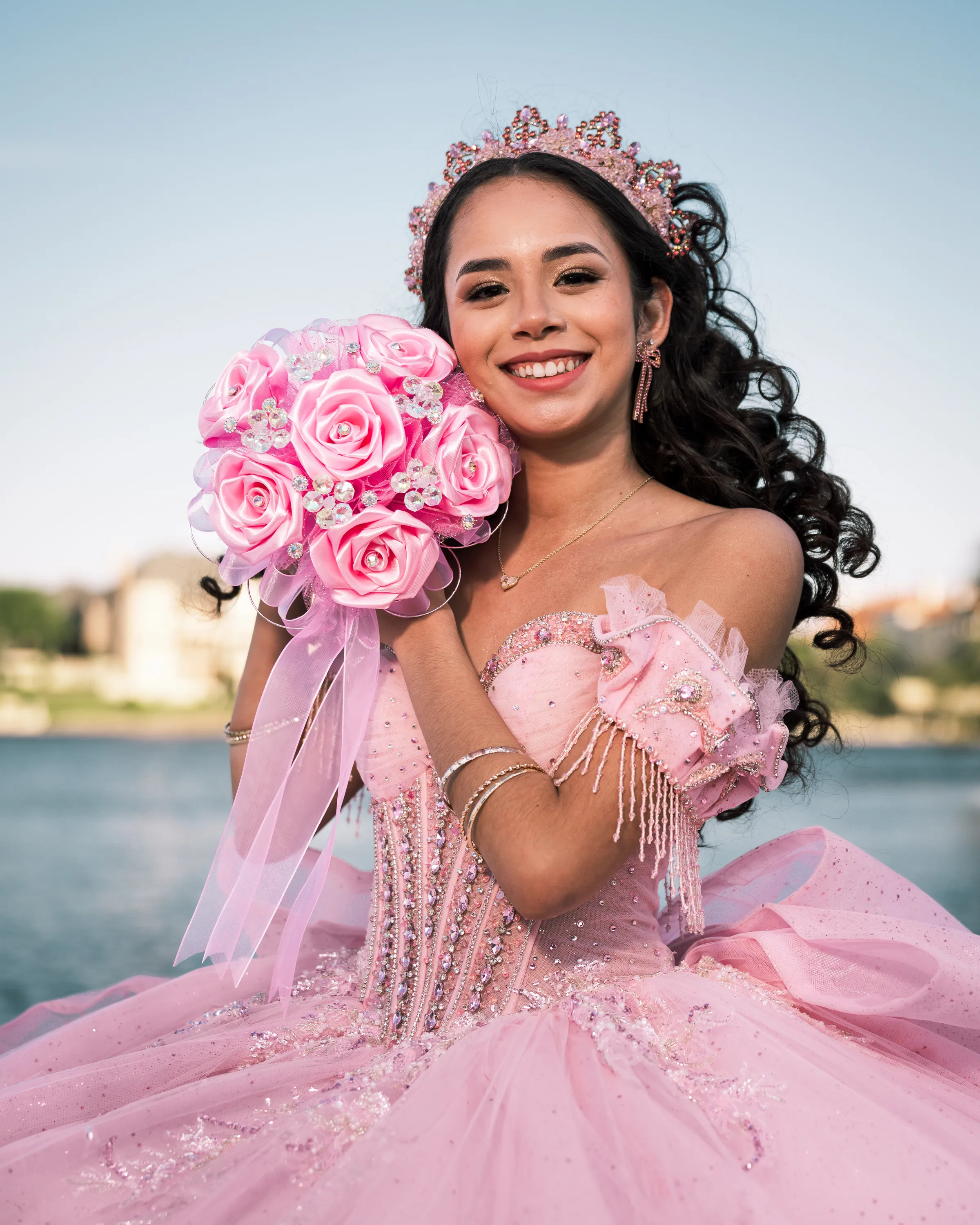 Quinceañera sonriente con vestido de quince rosa y corona sosteniendo un ramo de rosas rosadas junto al agua en Adriatica Village en McKinney, Texas