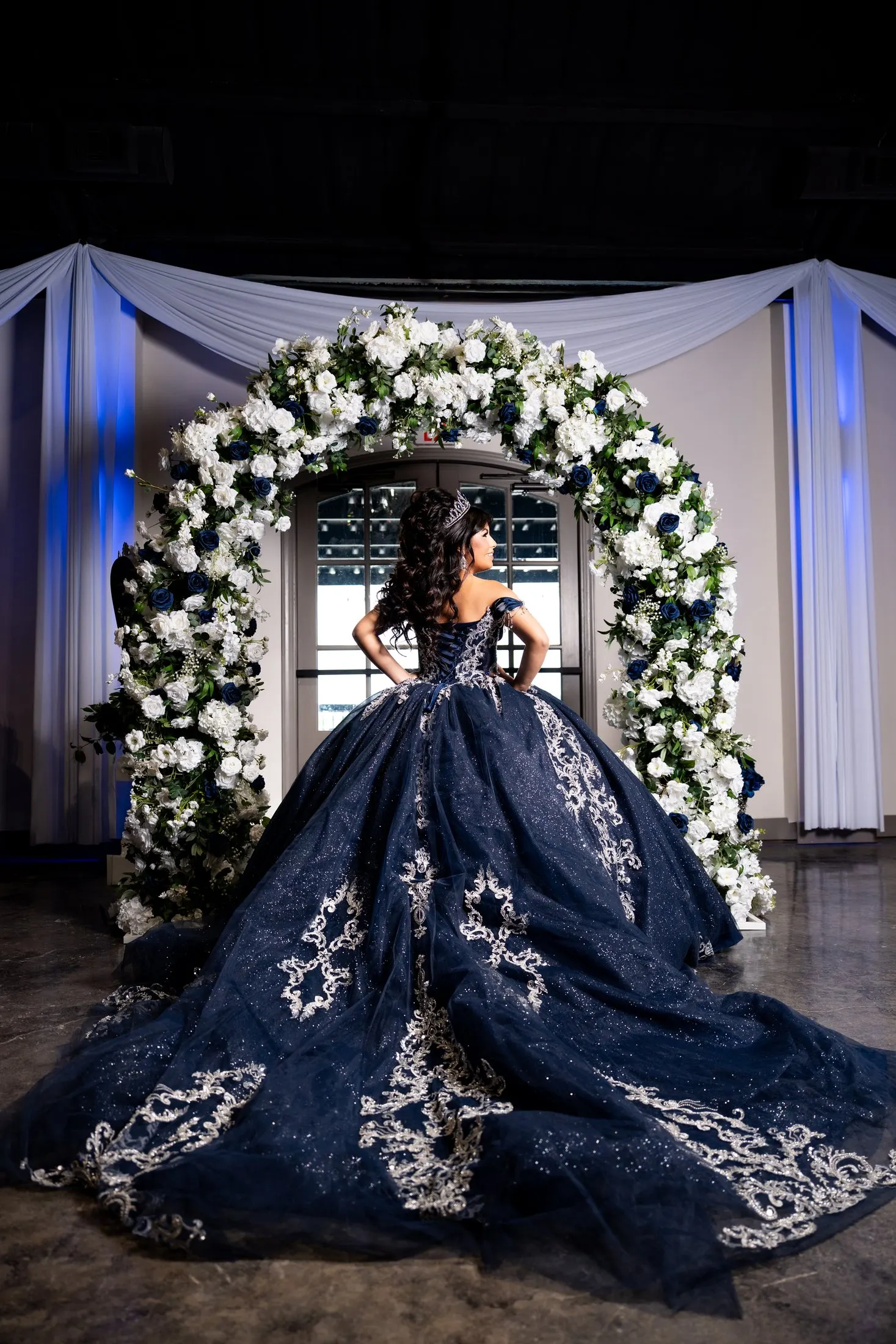 Julianna showing the train of her navy quince gown in front of a white flower arch at Diamond Ranch Event Center
