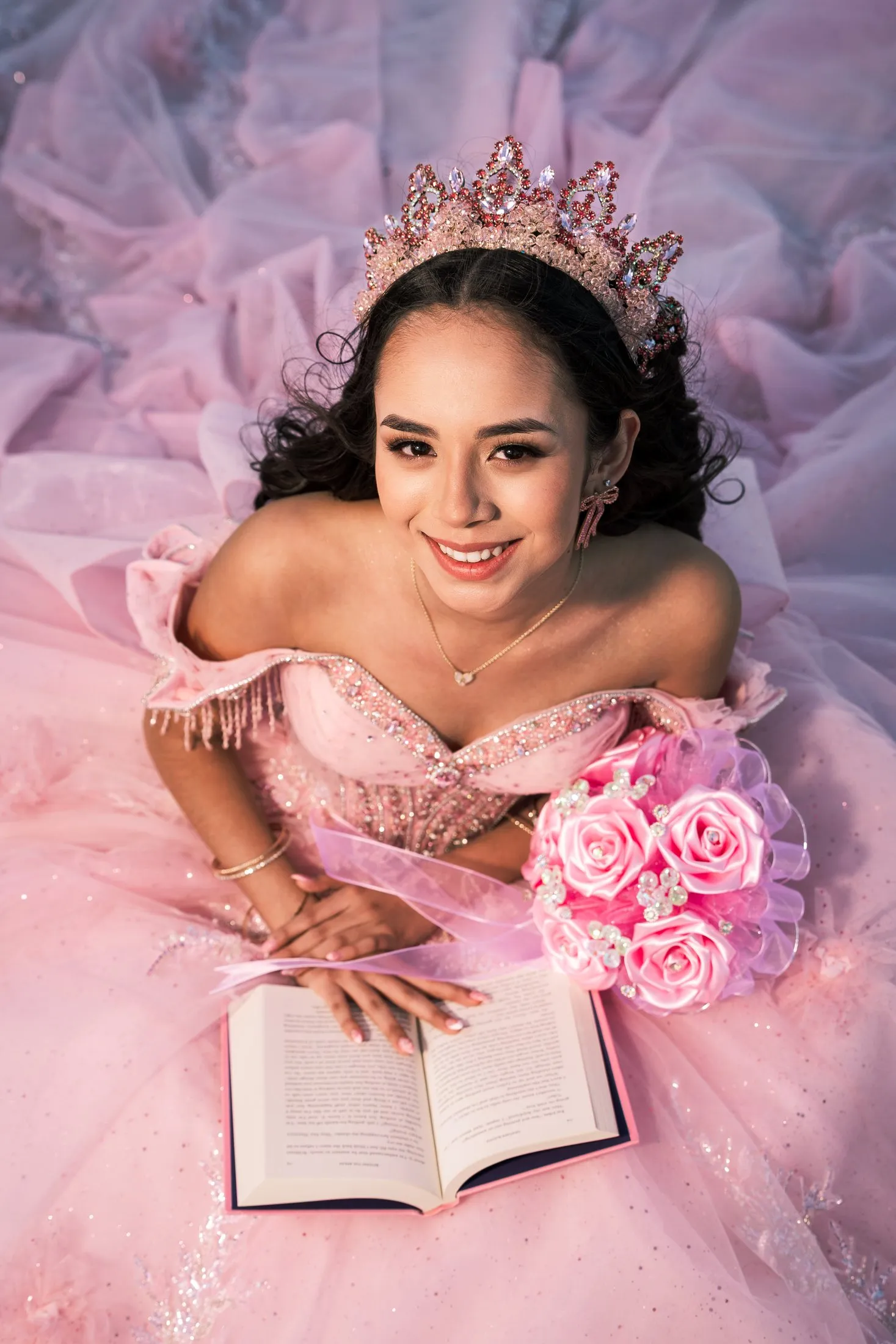 Overhead portrait of Rosalie in her pink quinceañera gown and rose-gold crown with an open book and pink rose bouquet