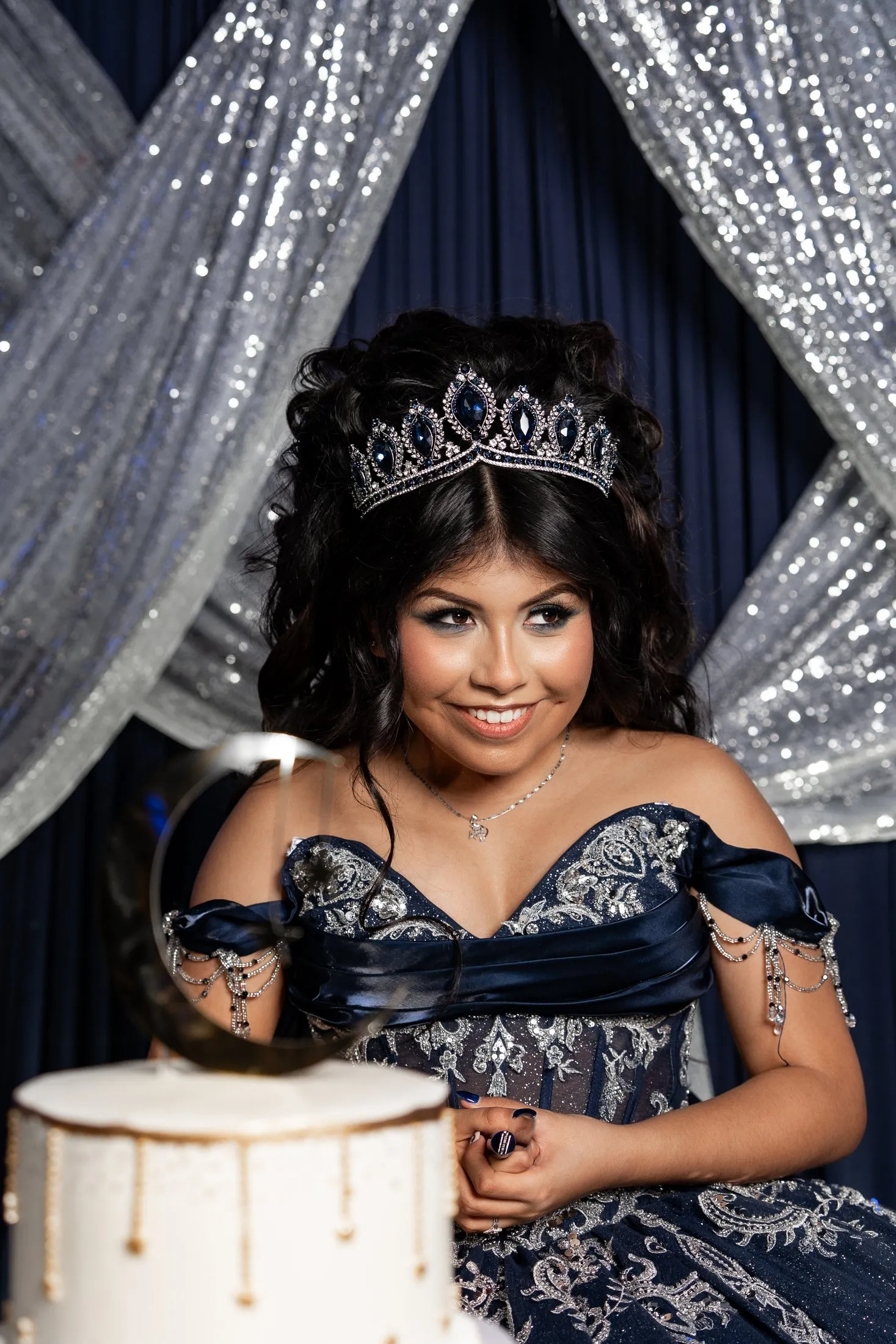 Quinceañera smiling by her cake in front of silver curtains
