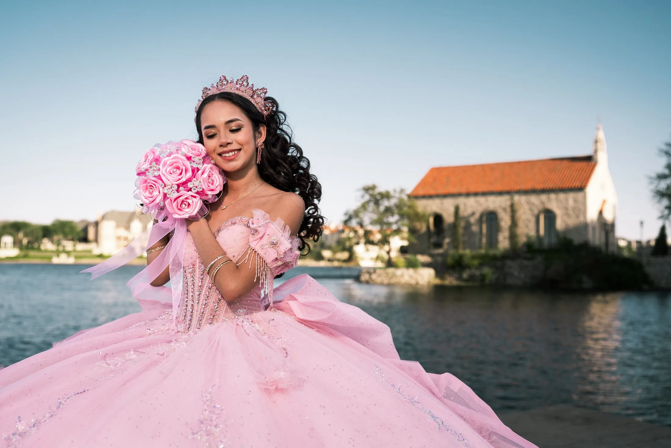Quinceañera hugging her pink bouquet beside the lakeside chapel in McKinney
